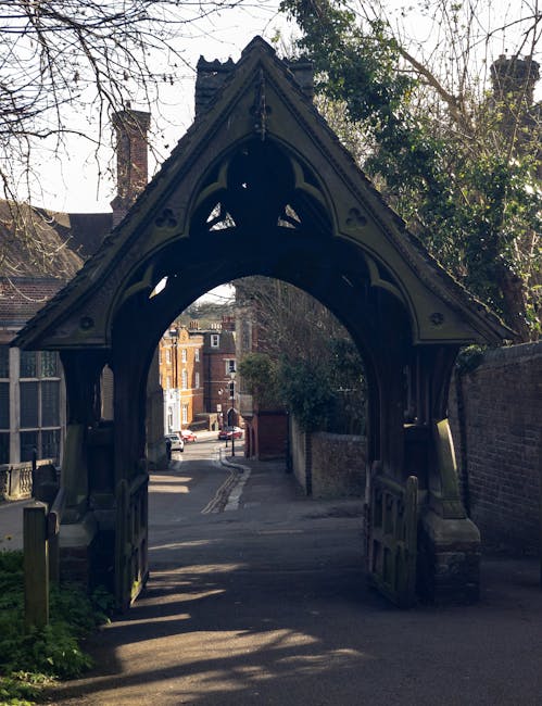 A narrow residential street viewed through an ornate, Gothic-style wooden archway with intricate carvings and a pointed arch top. The street beyond features a paved pathway flanked by brick buildings,