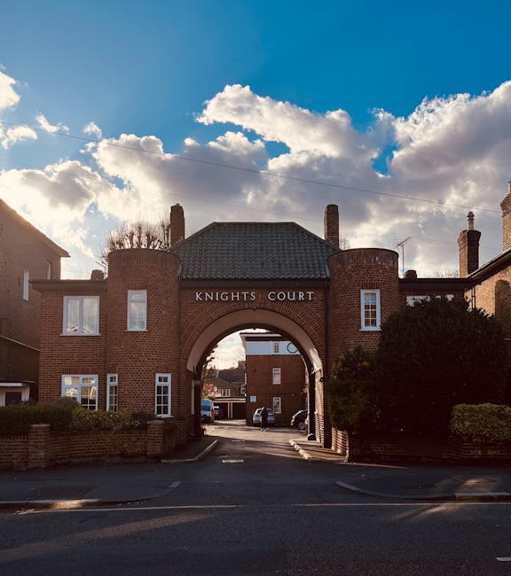 A narrow residential street viewed through an ornate, Gothic-style wooden archway with intricate carvings and a pointed arch top. The street beyond features a paved pathway flanked by brick buildings,