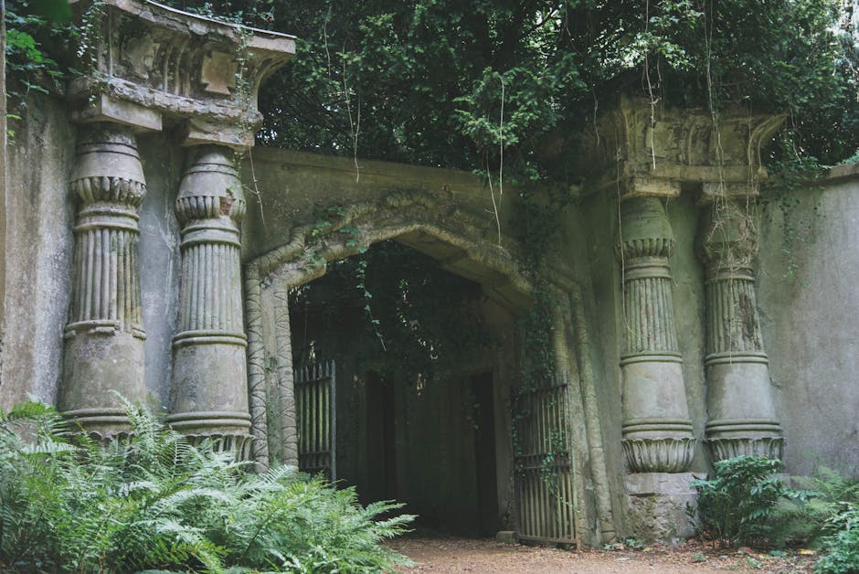 An ancient, overgrown stone archway with classical architectural elements, including fluted columns with ornate capitals and a curved entablature, serves as an entrance to a historic garden area. The 